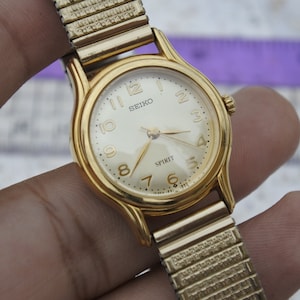 Könnte beinhalten: Eine goldfarbene Seiko Spirit Armbanduhr mit cremefarbenem Zifferblatt. Die Uhr hat goldene Zeiger und Stundenmarkierungen, mit dem Markennamen und "Spirit" auf dem Zifferblatt. Das Armband ist goldfarben und dehnbar.
