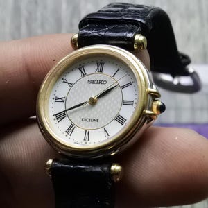 Könnte beinhalten: Eine goldfarbene Seiko Exceline Armbanduhr mit weißem Zifferblatt und schwarzem Lederarmband.