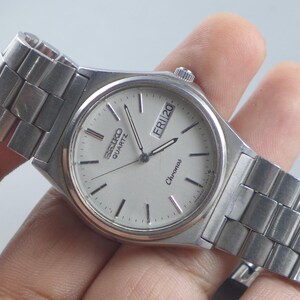 Könnte beinhalten: Eine silberne Seiko Quartz Chronos Uhr mit weißem Zifferblatt. Die Uhr zeigt Tag und Datum an. Die Uhr hat ein silbernes Armband und ein rundes Zifferblatt. Die Uhr wird in einer Hand gehalten.