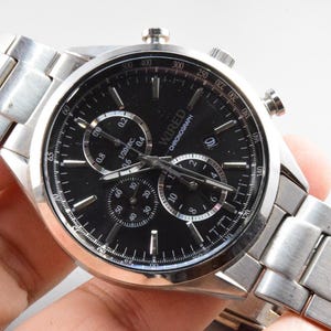May include: A silver-toned WIRED Chronograph watch with a black face. The watch features multiple sub-dials, a date display, and a silver band. The watch is held by a hand.
