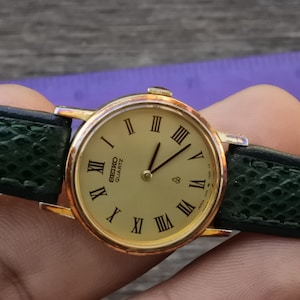 May include: A gold-tone Seiko Quartz watch with a green leather band. The watch face has Roman numeral hour markers and a gold-tone second hand.
