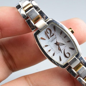 Könnte beinhalten: Eine Seiko Armbanduhr mit rechteckigem silberfarbenem Gehäuse und einem zweifarbigen Gliederarmband in Silber und Gold. Das Zifferblatt ist weiß mit gesprenkelter Textur, dunkelbraunen arabischen Ziffern auf 12 und 6 sowie dunkelbraunen Zeigern.