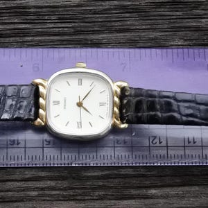 May include: A gold-tone and silver-tone wristwatch with a white face and black alligator-style band. The watch face has black Roman numeral hour markers and a gold-tone second hand.