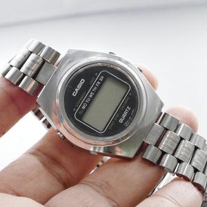 Puede incluir: Un reloj digital Casio plateado con pantalla negra y correa de acero inoxidable. La esfera muestra el día de la semana y la palabra "Quartz". El reloj está en una mano.