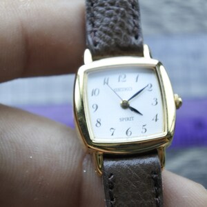 Könnte beinhalten: Eine goldfarbene Uhr mit quadratischem Zifferblatt und braunem Lederarmband. Das Zifferblatt ist weiß mit schwarzen Zahlen und Zeigern. Das Wort "Spirit" ist auf dem Zifferblatt aufgedruckt.