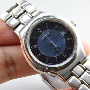 Op de afbeelding: Een zilverkleurig Citizen horloge met een donkerblauwe wijzerplaat en een zilveren band. De wijzerplaat heeft zwarte cijfers en het Citizen-logo. Het horloge wordt door een hand vastgehouden.