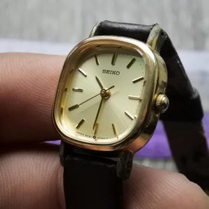 Könnte beinhalten: Eine goldfarbene Seiko Armbanduhr mit quadratischem Zifferblatt und schwarzem Lederarmband. Das Zifferblatt hat einen weißen Hintergrund mit schwarzen Stunden- und Minutenzeigern und schwarzen römischen Ziffern als Stundenmarkierungen.