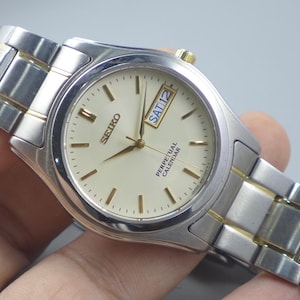 May include: A Seiko Perpetual Calendar watch with a silver and gold-tone stainless steel band. The watch face is cream-colored with gold-tone markers and hands. The day and date are displayed at the 3 o'clock position.