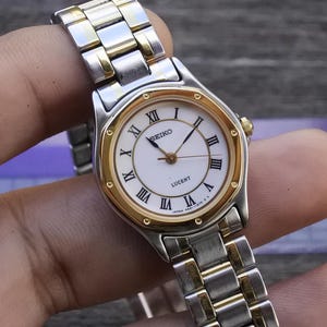 Puede incluir: Un reloj de pulsera Seiko Lucent con una correa de acero inoxidable plateada y dorada. La esfera del reloj es blanca con agujas doradas y números romanos. El reloj está sujeto por una mano.
