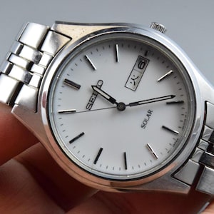 Pode incluir: Um relógio de pulso Seiko Solar em tom prateado com mostrador branco, marcadores de hora prateados e ponteiros prateados. O relógio possui uma janela de dia e data na posição de três horas com caracteres japoneses e pulseira de metal.