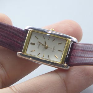 Puede incluir: Un reloj Seiko rectangular con un bisel dorado y una esfera blanca. El reloj tiene agujas y marcadores dorados. La correa del reloj es de color burdeos oscuro con un patrón texturizado. El reloj es de estilo clásico.
