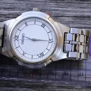 May include: A silver Seiko wristwatch with a white face and black Roman numeral hour markers. The watch has a silver metal band.