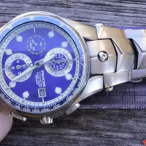 May include: A silver and blue chronograph wristwatch with a titanium band. The watch face features a blue background with white markings and a date window. The watch has a chronograph function and a tachymeter scale.