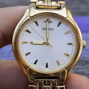 Könnte beinhalten: Eine goldfarbene Seiko Armbanduhr mit weißem Zifferblatt und schwarzen Zeigern. Die Uhr hat ein goldfarbenes Metallband und ein Datumsfenster bei 3 Uhr. Das Zifferblatt der Uhr hat ein strukturiertes Muster und die Worte "SEIKO" und "JAPAN V701" sind auf dem Zifferblatt aufgedruckt.
