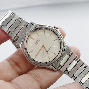 Könnte beinhalten: Eine silberfarbene Seiko Quartz-Uhr mit cremefarbenem Zifferblatt und goldenen Zeigern. Die Uhr hat ein rechteckiges Gliederarmband. Das Zifferblatt zeigt das Seiko Quartz-Logo und das Wort "chronos".