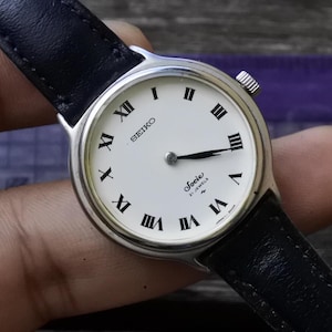 May include: A silver Seiko Socie 21 Jewels wristwatch with a white face and black leather band. The watch has Roman numeral hour markers and a black second hand.
