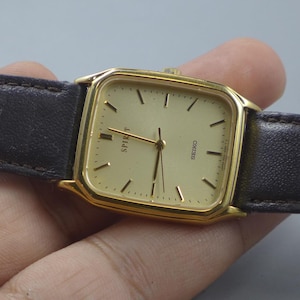 Könnte beinhalten: Eine goldfarbene Seiko Spirit Uhr mit rechteckigem Zifferblatt und dunkelbraunem Lederarmband. Das Zifferblatt ist hellgoldfarben mit schwarzen Stundenmarkierungen und Zeigern. Das Wort "SPIRIT" ist auf dem Zifferblatt aufgedruckt.