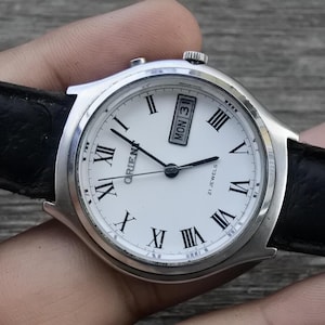 May include: A silver wristwatch with a white face and black leather band. The watch face has Roman numeral hour markers, a date window, and the text "Orient 21 Jewels".