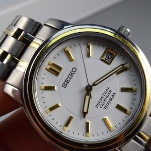 May include: A Seiko Perpetual Calendar Titanium wristwatch featuring a white dial with gold-tone hour markers and hands. The watch has a two-tone silver and gold metal link bracelet and a date window positioned at the three o'clock mark on the face.
