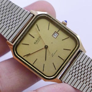 Puede incluir: Un reloj Rado de cuarzo dorado con una esfera cuadrada y una correa de malla plateada. La esfera del reloj es dorada con agujas negras y marcadores de hora romanos negros. El reloj también tiene una ventana de fecha en la posición de las 3 en punto.