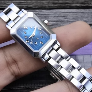 Könnte beinhalten: Eine silberne Uhr mit blauem Zifferblatt und einem silbernen Metallband. Das Zifferblatt der Uhr hat ein Sternmuster und silberne Zeiger.