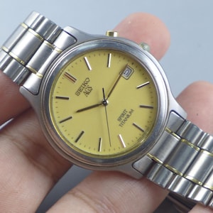 Könnte beinhalten: Eine silberfarbene Seiko AGS Spirit Titanium Uhr mit gelbem Zifferblatt. Die Uhr verfügt über eine Datumsanzeige und ein silber-goldfarbenes Metallarmband. Die Uhr wird in einer Hand gehalten.