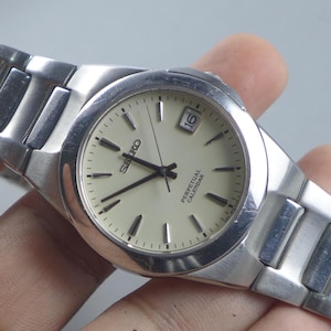 May include: A silver-toned Seiko watch with a cream-colored face. The watch features black hands and markers, a date display at the 3 o'clock position, and a stainless steel band. The watch face reads "SEIKO PERPETUAL CALENDAR".