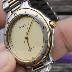 Könnte beinhalten: Eine silberne und goldene zweifarbige Armbanduhr mit weißem Zifferblatt und schwarzen römischen Ziffern. Auf dem Zifferblatt sind die Wörter "DOLCE" und "SEIKO" aufgedruckt.