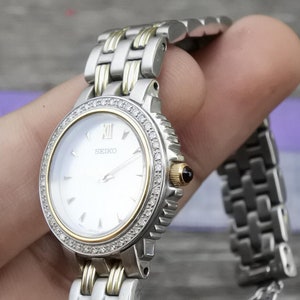 May include: A silver and gold two-tone watch with a white face and diamond accents around the bezel. The watch face has a silver-tone 'SEIKO' logo and Roman numeral hour markers.