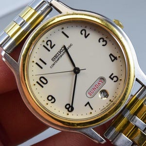 Pode incluir: Um relógio de pulso Seiko Contemporary Basic com mostrador redondo na cor creme, algarismos arábicos pretos e ponteiros pretos. O relógio possui uma moldura dourada, uma caixa prateada e uma pulseira de elos metálicos em tons de ouro e prata.