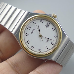 Puede incluir: Un reloj de pulsera Seiko Spirit con una correa de metal plateado y una esfera blanca. El reloj tiene un bisel y agujas doradas, con números negros y la fecha a las 3 en punto. El reloj es sostenido por una mano.