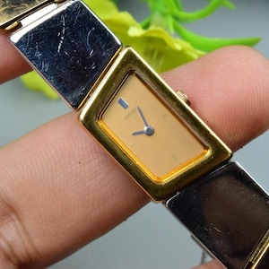 Puede incluir: Un reloj de pulsera dorado y plateado con una esfera rectangular. La esfera del reloj es de color beige claro con agujas azules y el nombre de la marca. La correa del reloj es una combinación de eslabones metálicos dorados y plateados.