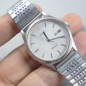 Pode incluir: Um relógio de pulso Seiko Chronos prateado com mostrador branco e exibição de data. O relógio tem uma pulseira prateada e é segurado por uma mão. O mostrador tem marcadores e ponteiros pretos.