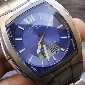 May include: A silver Casio Wave Ceptor Tough Solar watch with a blue face and a digital display. The watch has a black band and a silver case. The watch face has the words "Wave Ceptor Tough Solar" and "World Time Alarm DST" printed on it.