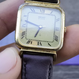 May include: A gold-toned square-shaped wristwatch with a black leather band. The watch face is white with black Roman numeral hour markers and black hands. The watch face also has the text "SEIKO QUARTZ" printed on it.