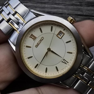 May include: A silver and gold two-tone Seiko wristwatch with a white face and black hands. The watch has Roman numeral hour markers and a date window at the 3 o'clock position. The watch is on a silver and gold two-tone metal band.