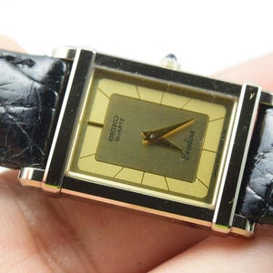 May include: A rectangular Seiko Quartz Exceline wristwatch featuring a gold-tone dial with thin black hour markers and gold hands. The watch has a polished silver-tone metal case and is attached to a black textured leather strap with a glossy finish.