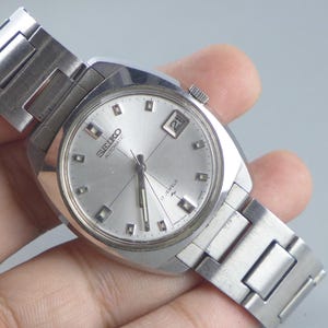 Pode incluir: Relógio de pulso automático Seiko prateado com mostrador retangular e visor de data. O relógio tem um mostrador prateado com marcadores de hora retangulares e uma pulseira de aço inoxidável. O diâmetro do mostrador é de cerca de 3,5 cm.