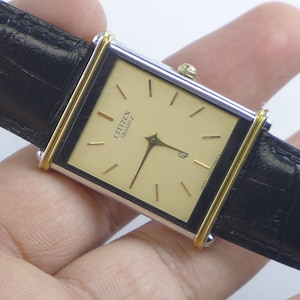 Puede incluir: Un reloj Citizen Quartz rectangular con esfera y agujas doradas, en una caja plateada y dorada. El reloj tiene una correa de cuero negro. La esfera del reloj muestra el logotipo de Citizen Quartz.