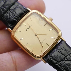 May include: A gold-tone Rado watch with a square face and black leather strap. The watch face has gold-tone hands and markers, and the word "RADO" is printed in gold on the face. The watch also has the word "QUARTZ" printed on the face.