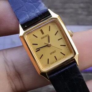 Könnte beinhalten: Eine goldfarbene Seiko Spirit Armbanduhr mit quadratischem Zifferblatt und schwarzem Lederarmband. Das Zifferblatt ist hellgoldfarben mit schwarzen Stundenmarkierungen und Zeigern. Die Wörter "SEIKO" und "SPIRIT" sind auf dem Zifferblatt aufgedruckt.