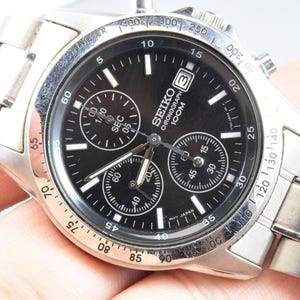 May include: A silver-tone stainless steel Seiko chronograph watch with a black dial, three sub-dials, and a date window at the three o'clock position. The watch features a tachymeter bezel, luminous hour markers, and a metal link bracelet strap.