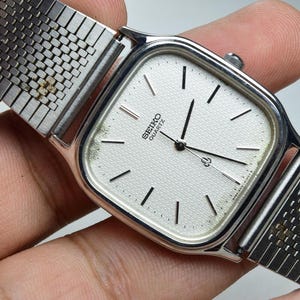 Op de afbeelding: Een zilverkleurig Seiko Quartz polshorloge met een vierkante kast met afgeronde hoeken en een getextureerde gebroken witte wijzerplaat. Het horloge heeft een metalen schakelarmband en zwarte uurmarkeringen met bijpassende zwarte wijzers.