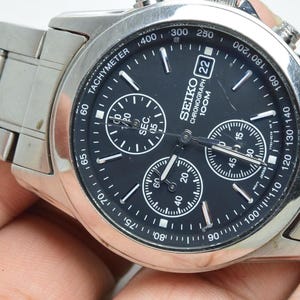 May include: A silver-tone stainless steel Seiko chronograph watch with a black dial, three sub-dials, a tachymeter scale on the bezel, and a date window at the three o'clock position. The watch features a metal link bracelet and white markers.