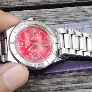 Könnte beinhalten: Eine silberne Armbanduhr mit rotem Zifferblatt und drei Hilfszifferblättern. Die Uhr hat ein silbernes Metallband.