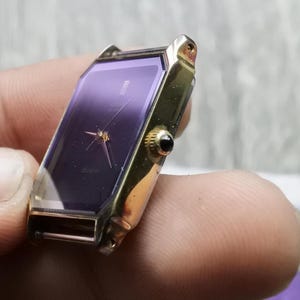 Puede incluir: Un reloj de pulsera dorado con una esfera cuadrada de color morado y agujas doradas. El reloj está en la muñeca de una persona.