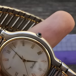 May include: A silver and gold two-tone Seiko Dolce watch with a white face and Roman numeral hour markers. The watch has a silver band with gold accents.