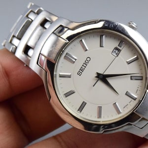 Könnte beinhalten: Eine silberfarbene Seiko Armbanduhr mit rundem weißem Zifferblatt, silberfarbenen Stundenmarkierungen, schwarzen Zeigern und einer Datumsanzeige auf der Drei-Uhr-Position. Die Uhr hat ein Gliederarmband aus Edelstahl mit poliertem Finish.