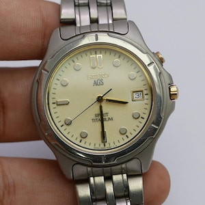 Könnte beinhalten: Eine Seiko AGS Spirit Titanium Armbanduhr mit silberfarbenem Titanarmband und -gehäuse. Das Zifferblatt der Uhr ist hellcremefarben mit goldfarbenen Akzenten, einschließlich der Zeiger und Stundenmarkierungen. Das Datum wird auf 3 Uhr angezeigt.