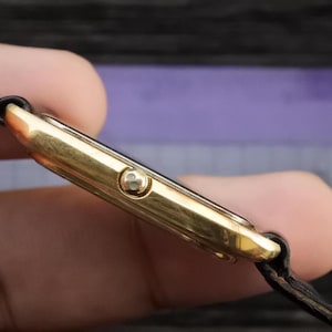 Könnte beinhalten: Eine goldfarbene Uhr mit einem schwarzen Lederarmband. Die Uhr hat ein quadratisches Zifferblatt und ein schlichtes Design.
